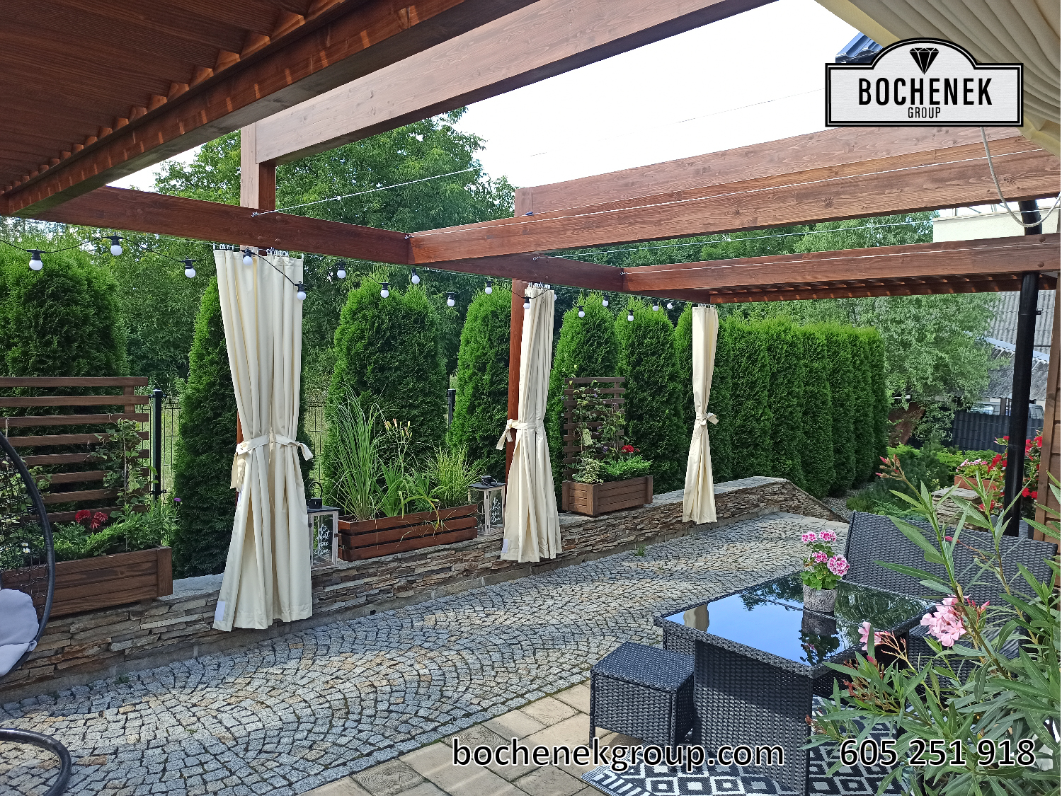 Pergola Kleopatra na drewnianym tarasie z meblami ogrodowymi, parasolami i zadbanym trawnikiem